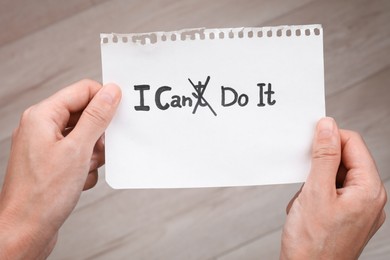 Photo of Motivation concept. Woman holding paper with changed phrase from I Can't Do It into I Can Do It by crossing over letter T indoors, closeup