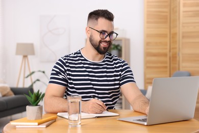 Young man writing in notebook while working on laptop at wooden table indoors