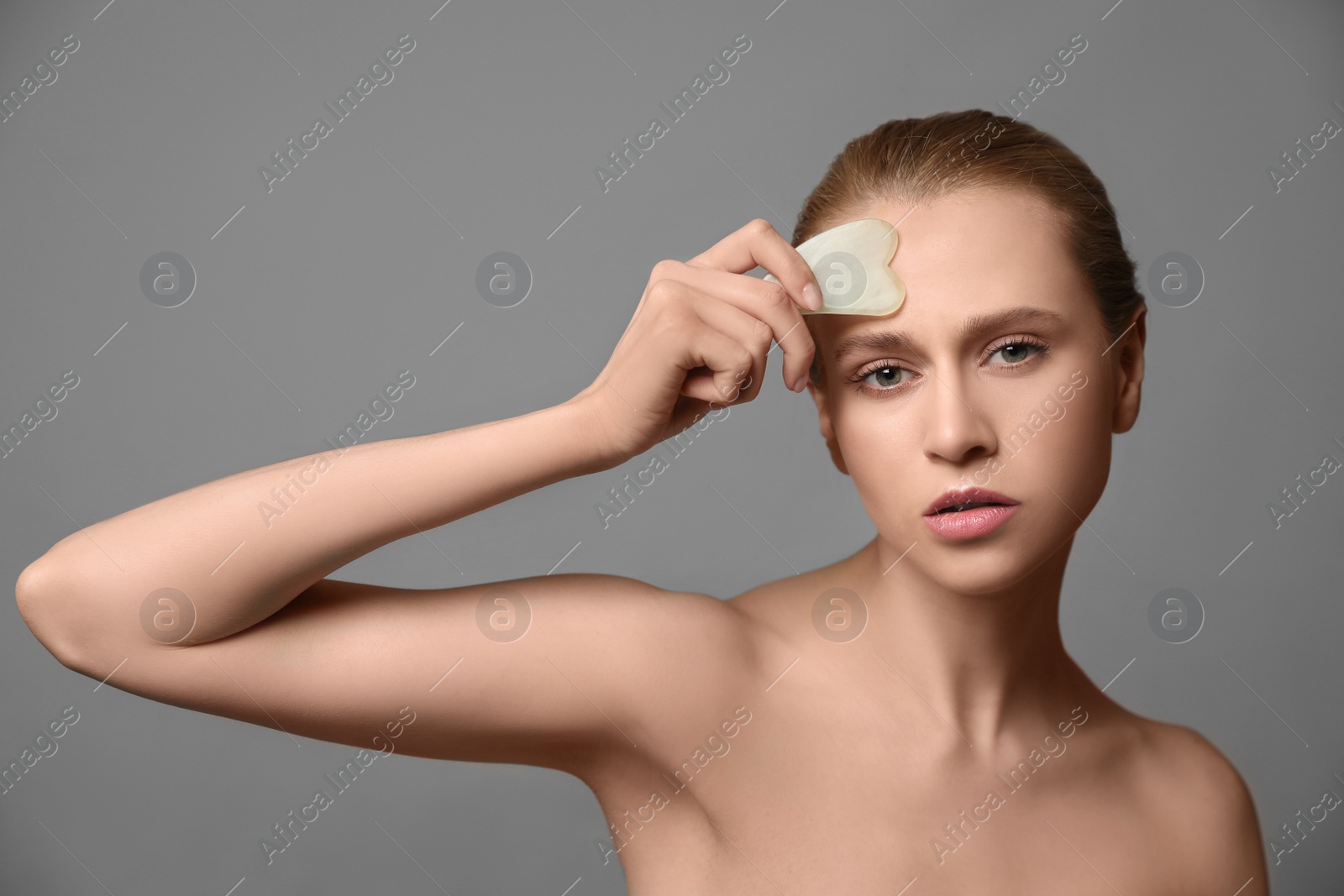 Photo of Beautiful young woman doing facial massage with gua sha tool on grey background