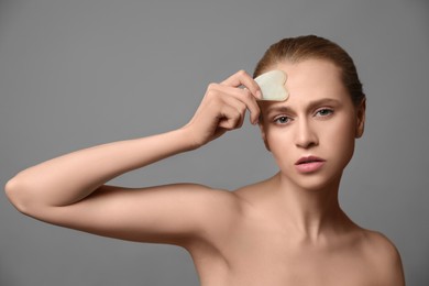 Photo of Beautiful young woman doing facial massage with gua sha tool on grey background