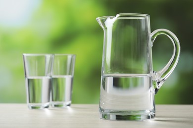 Photo of Jug and glasses with clear water on white table against blurred green background, closeup