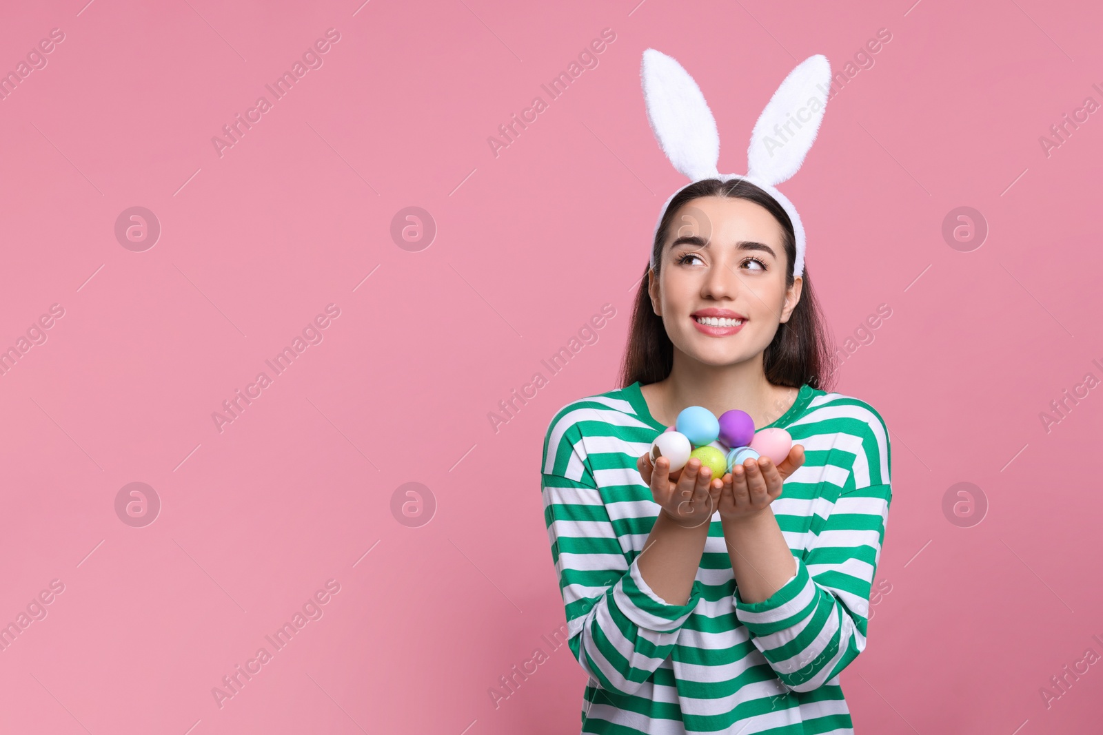 Photo of Happy woman in bunny ears headband holding painted Easter eggs on pink background. Space for text.