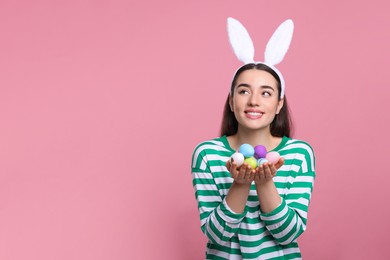 Happy woman in bunny ears headband holding painted Easter eggs on pink background. Space for text.