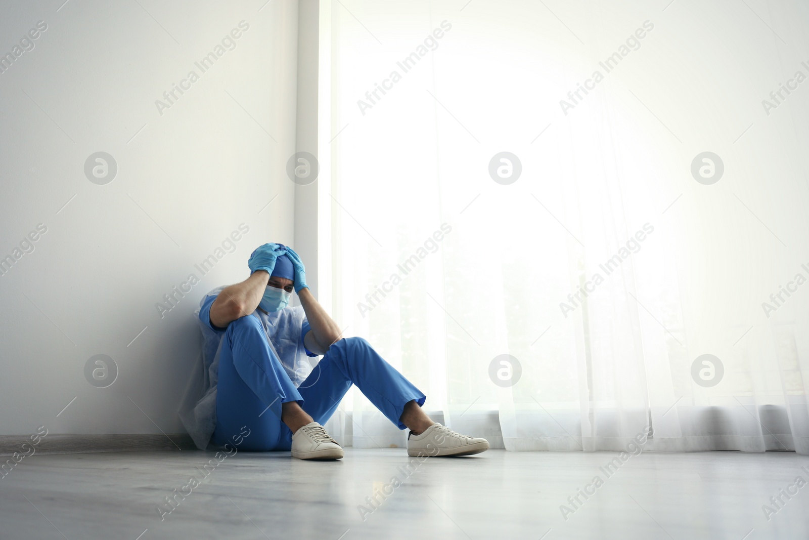 Photo of Exhausted doctor sitting on floor indoors, space for text. Stress of health care workers during COVID-19 pandemic