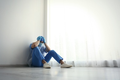 Photo of Exhausted doctor sitting on floor indoors, space for text. Stress of health care workers during COVID-19 pandemic