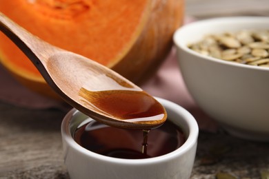 Photo of Wooden spoon with pumpkin oil over bowl and seeds on table, closeup