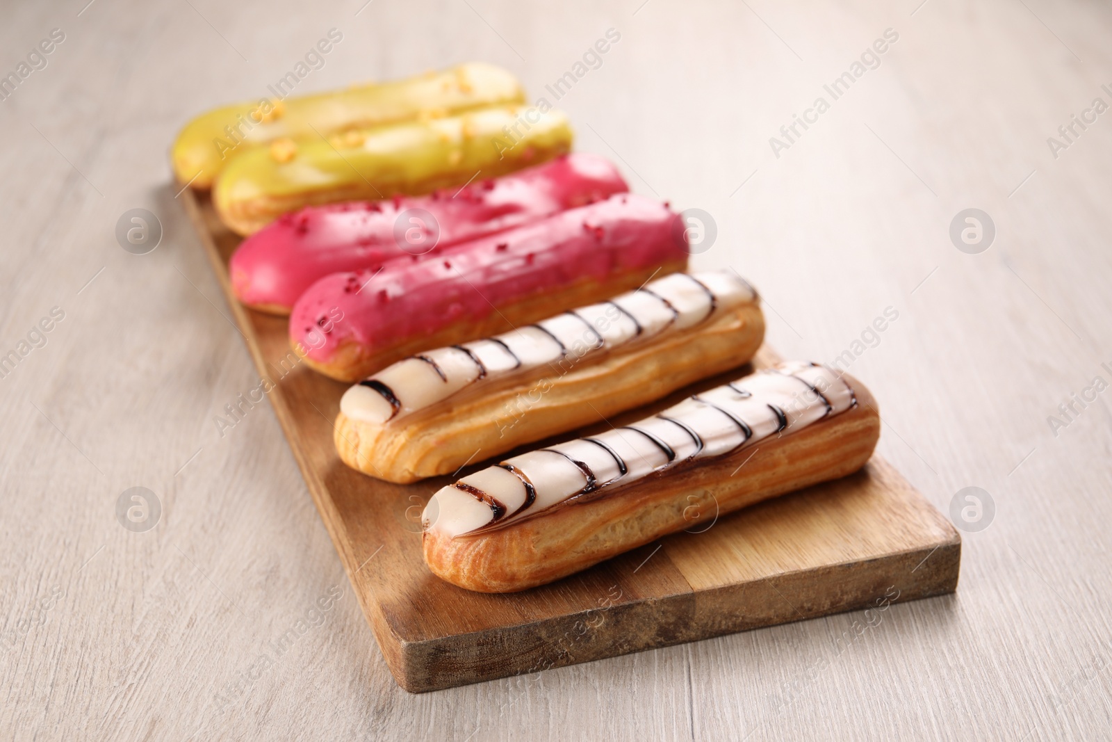 Photo of Board with different tasty glazed eclairs on light wooden table, closeup