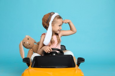 Photo of Cute little girl with toy bunny driving children's car on light blue background