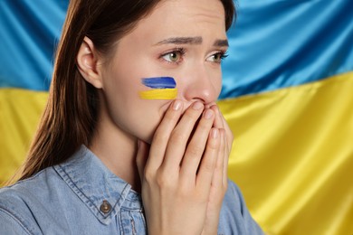 Photo of Sad young woman with clasped hands near Ukrainian flag, closeup. Space for text