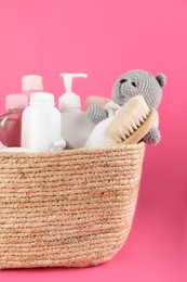 Wicker basket full of different baby cosmetic products, hairbrush and toy on pink background, closeup