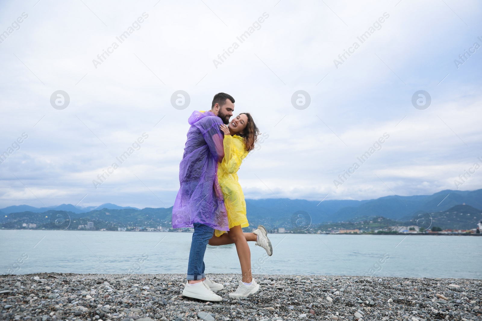 Photo of Young couple in raincoats enjoying time together under rain on beach, space for text
