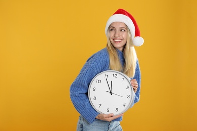 Photo of Woman in Santa hat with clock on yellow background, space for text. New Year countdown