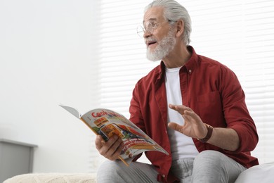Photo of Senior man in eyeglasses reading magazine on bed at home