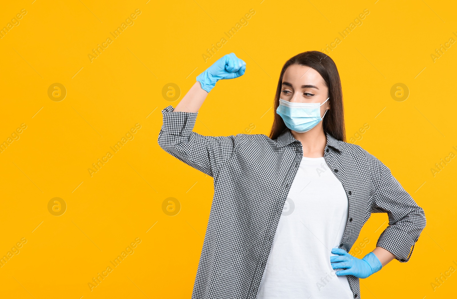 Photo of Woman with protective mask and gloves showing muscles on yellow background, space for text. Strong immunity concept