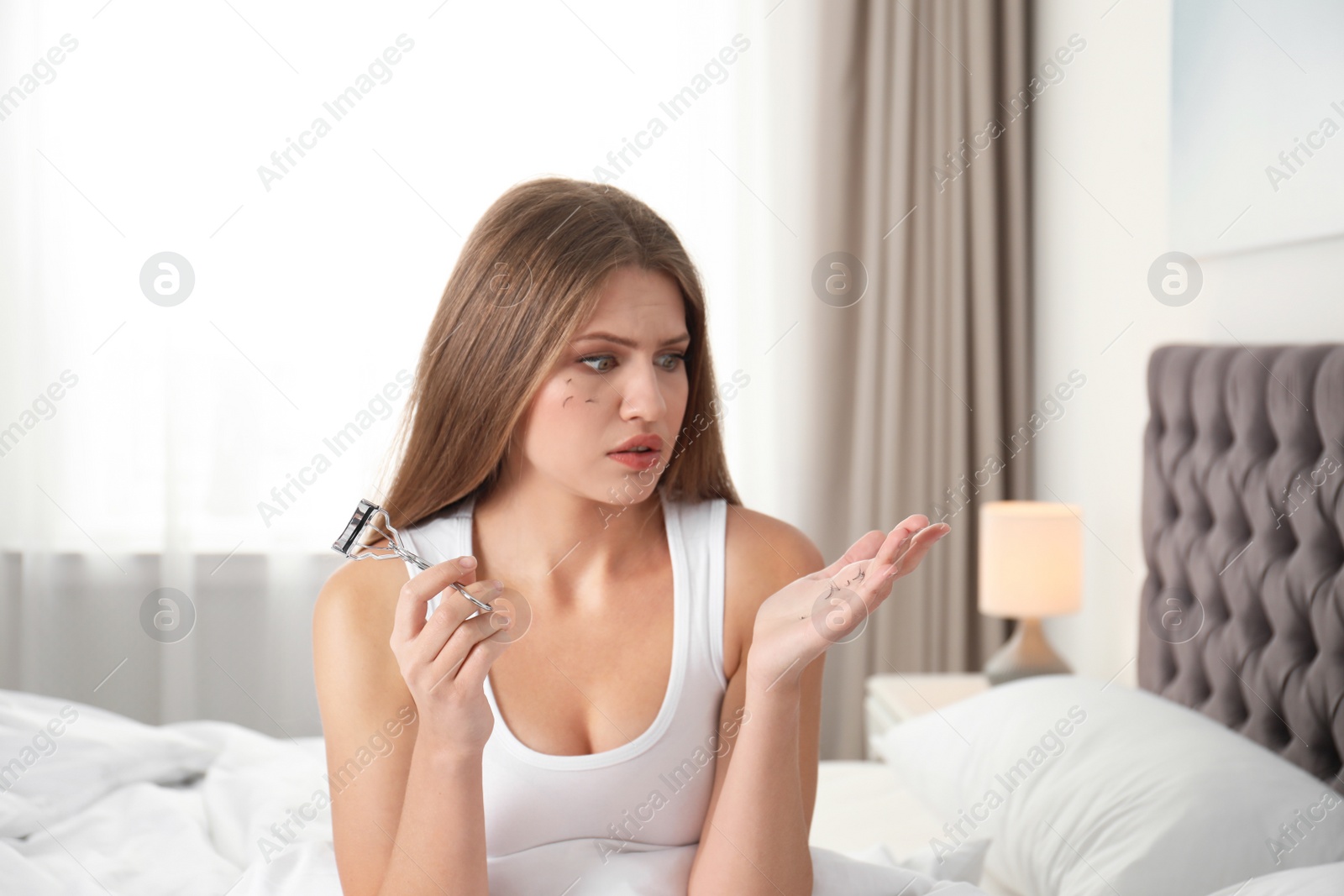 Photo of Emotional young woman holding fallen eyelashes and curler indoors