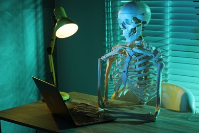 Photo of Waiting concept. Human skeleton sitting at wooden table with laptop indoors