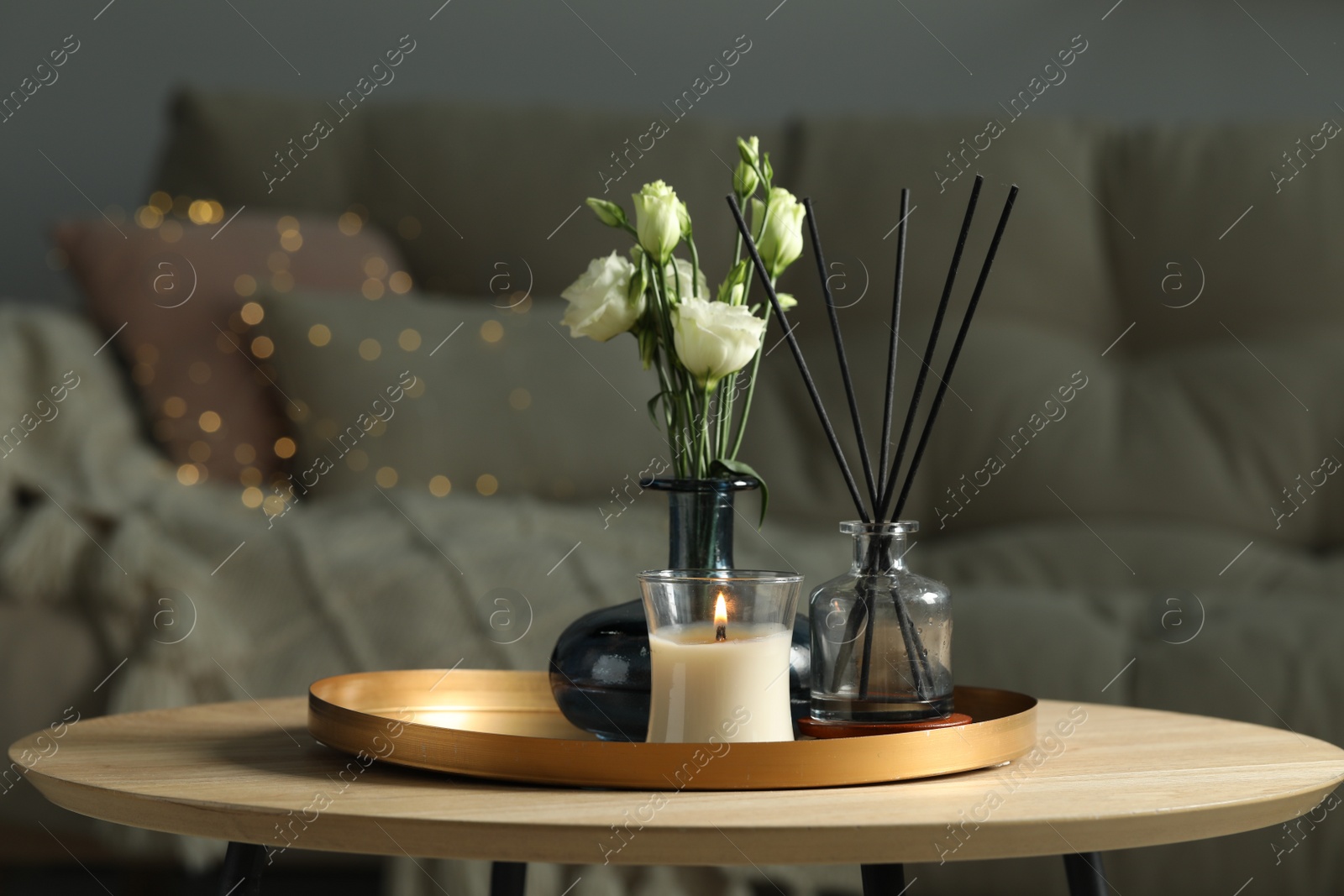 Photo of Burning candle with oil reed diffuser and bouquet on wooden table in living room