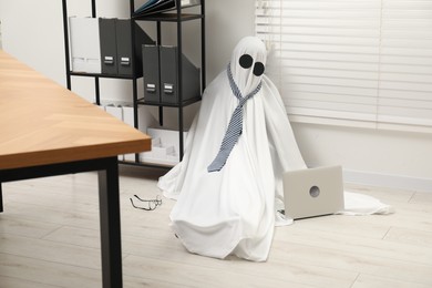 Photo of Overworked ghost. Man in white sheet with laptop on floor in office