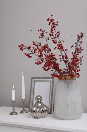 Photo of Hawthorn branches with red berries in vase, candles and frame on table indoors