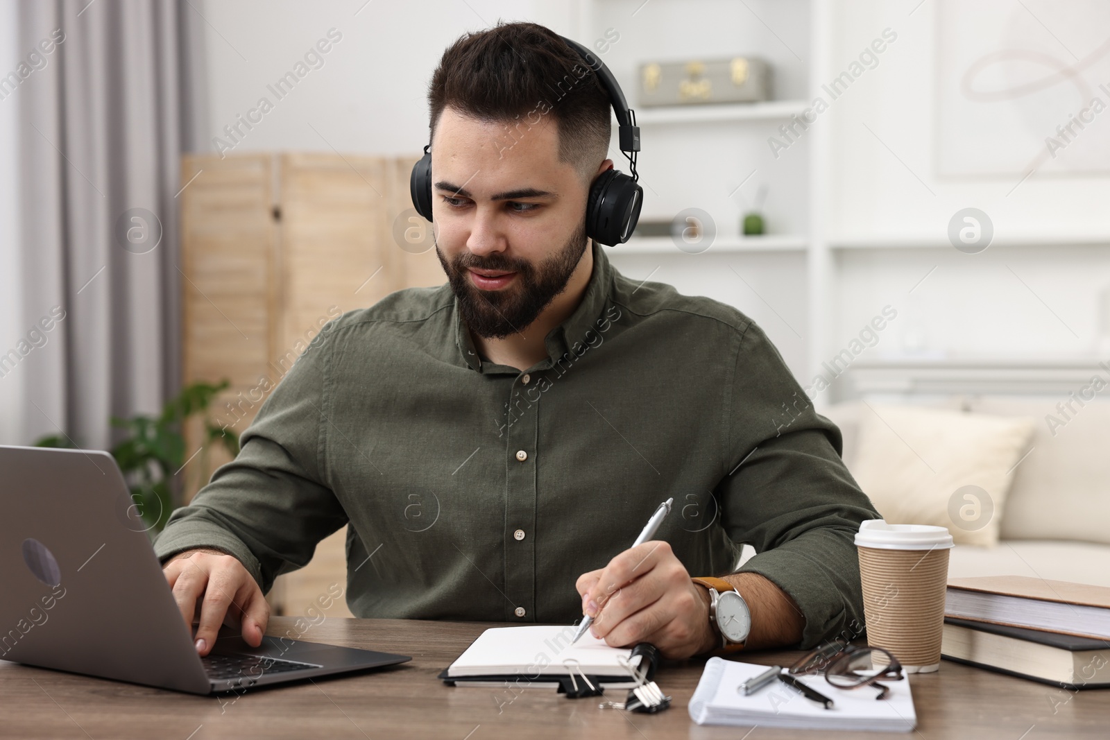 Photo of E-learning. Young man taking notes during online lesson at wooden table indoors