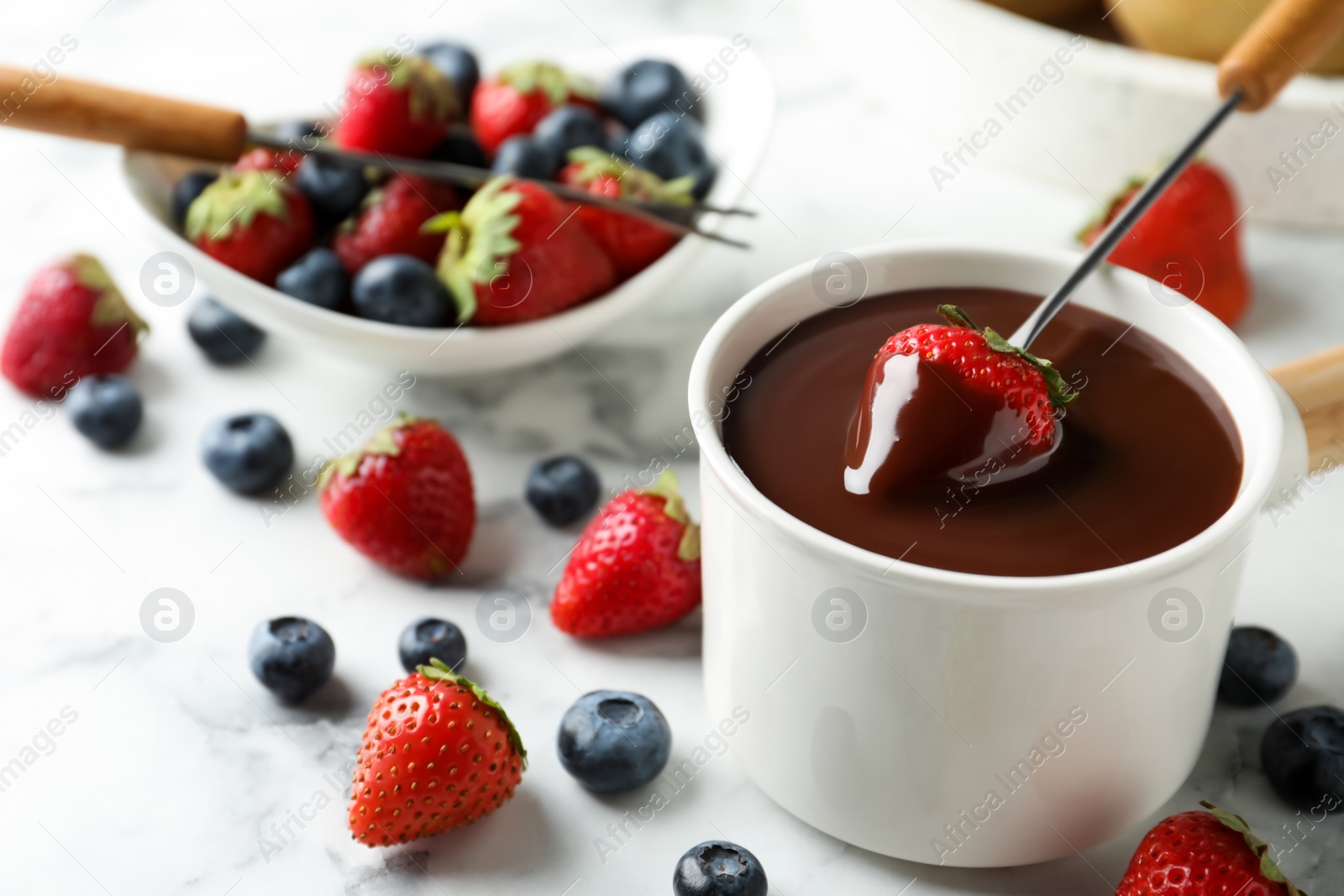 Photo of Dipping strawberry into fondue pot with chocolate on white marble table