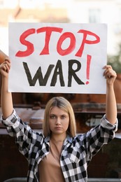Photo of Sad woman holding poster with words Stop War near broken tank in city
