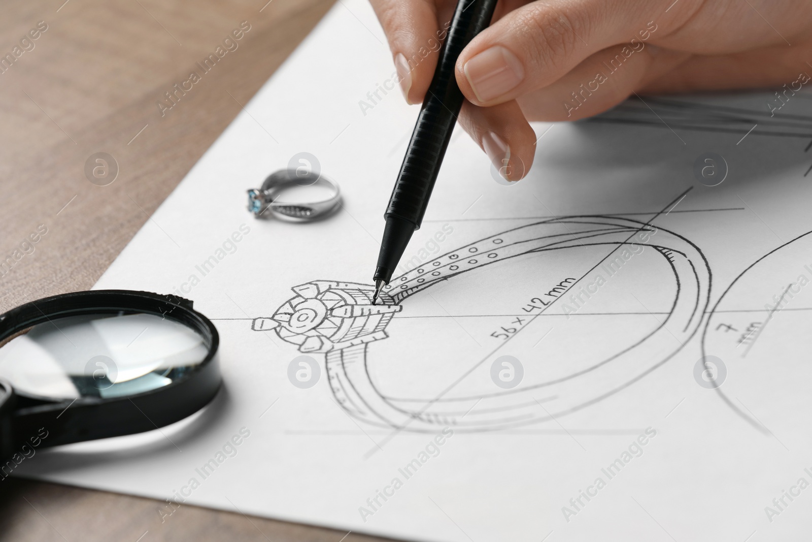 Photo of Jeweler drawing sketch of elegant ring on paper at wooden table, closeup