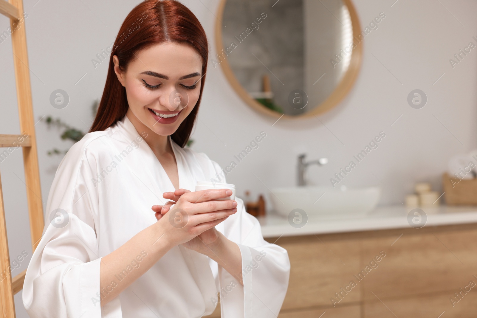 Photo of Beautiful young woman applying body cream onto hands in bathroom, space for text