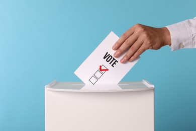 Man putting paper with word Vote and tick into ballot box on light blue background