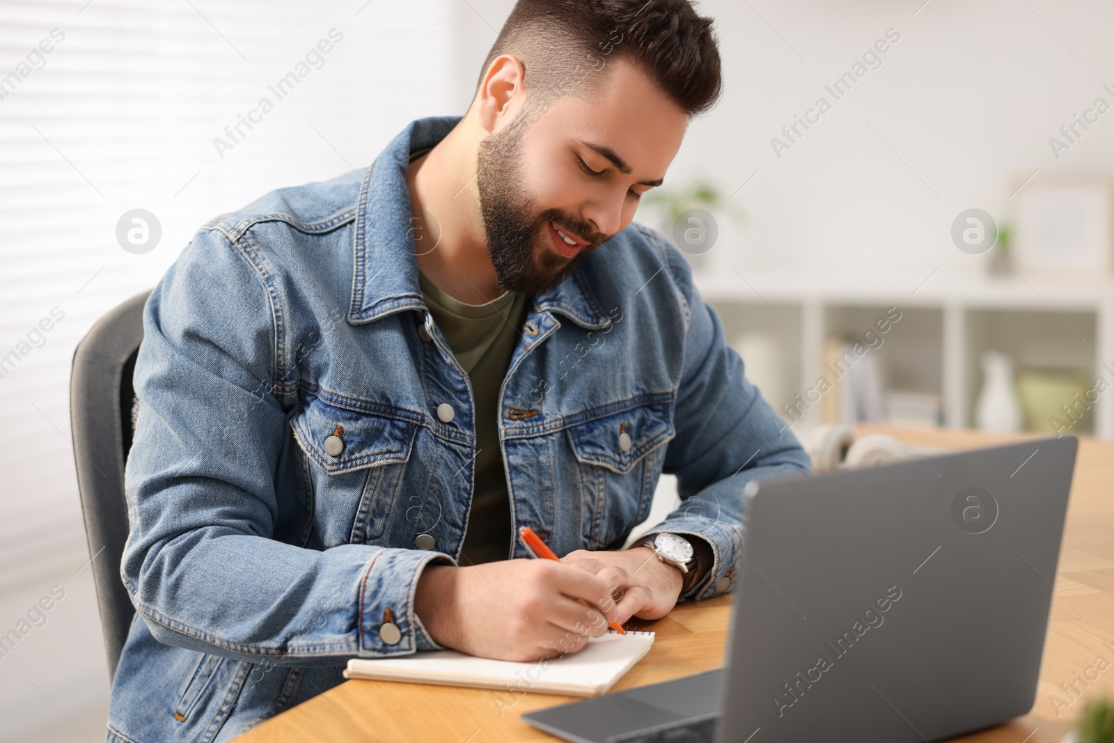 Photo of Young man writing down notes during webinar at table in room