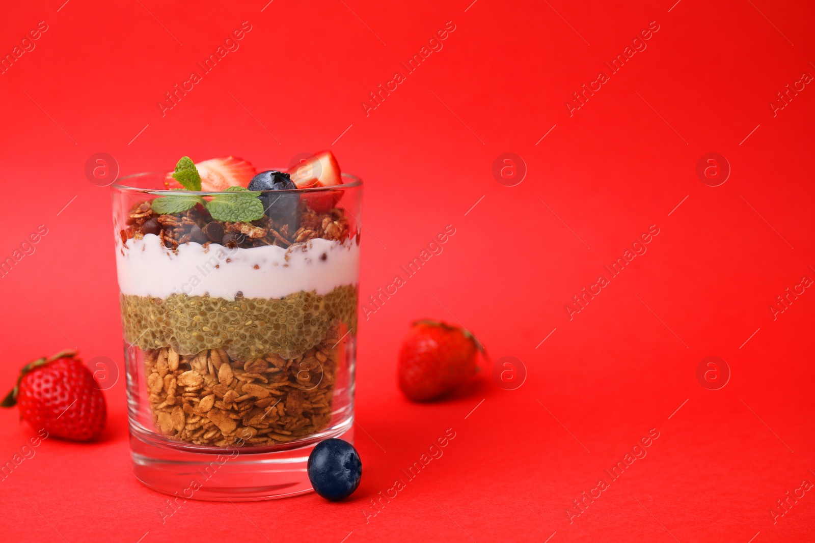 Photo of Tasty granola with berries, yogurt and chia seeds in glass on red background, space for text