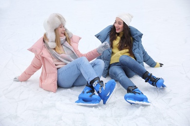 Happy women with figure skates sitting on ice rink outdoors