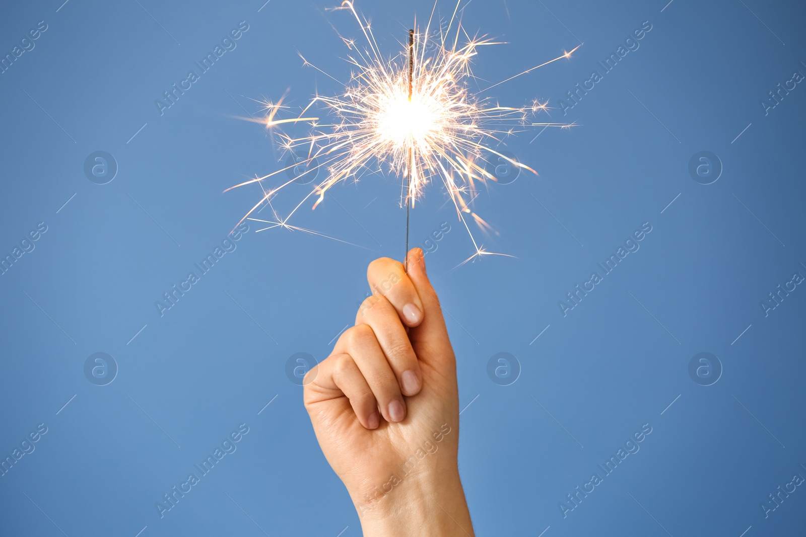 Photo of Woman holding bright burning sparkler on light blue background, closeup