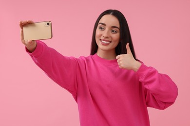 Photo of Smiling young woman taking selfie with smartphone and showing thumbs up on pink background, space for text