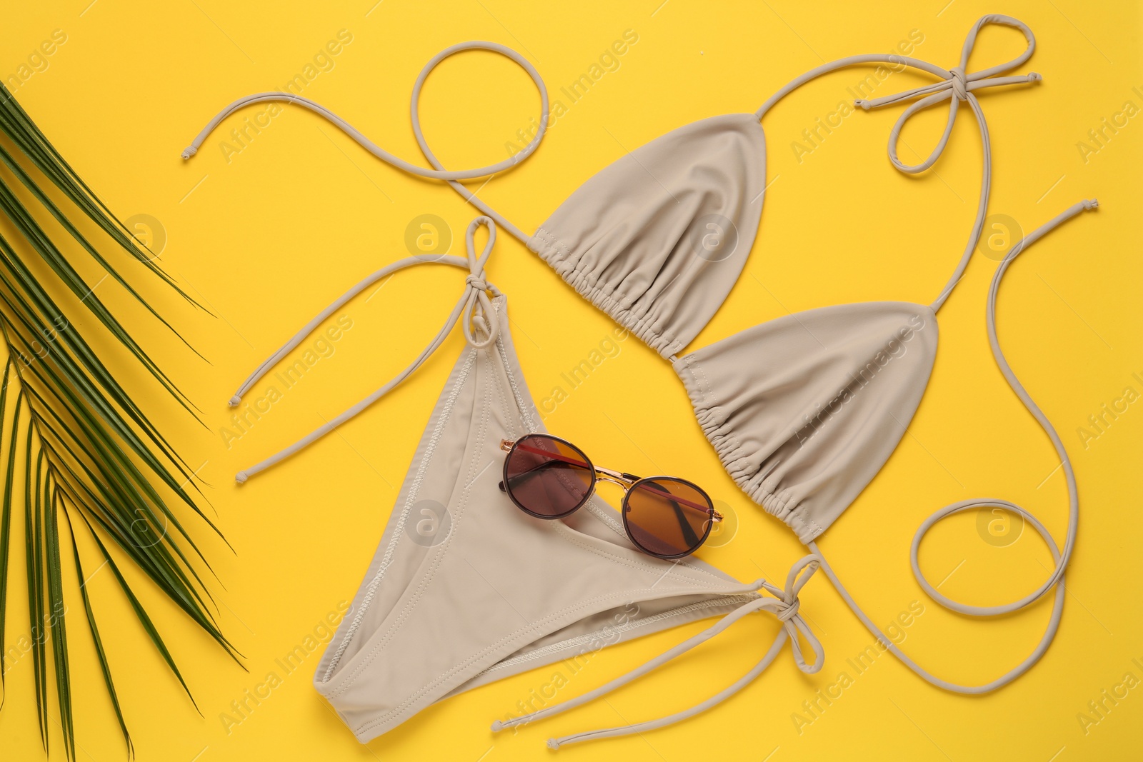 Photo of Stylish beige bikini, sunglasses and leaf on yellow background, flat lay