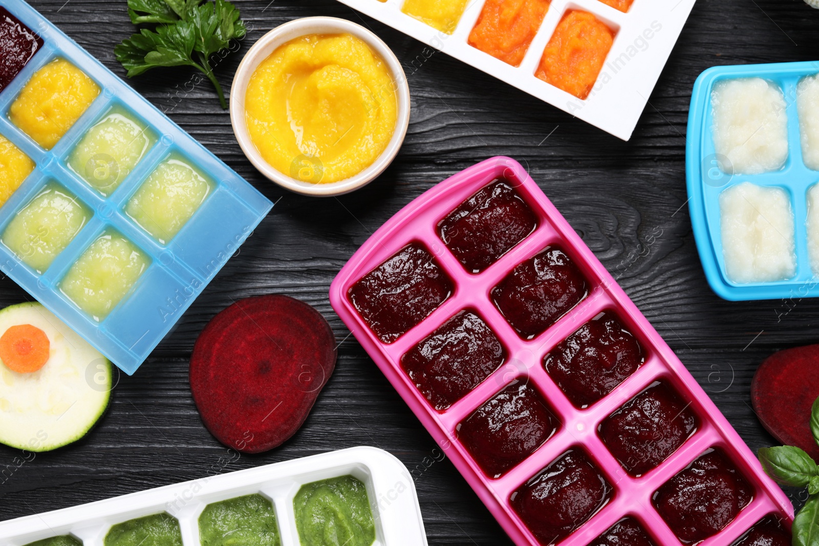 Photo of Different frozen purees in ice cube trays and ingredients of black wooden table, flat lay. Ready for freezing