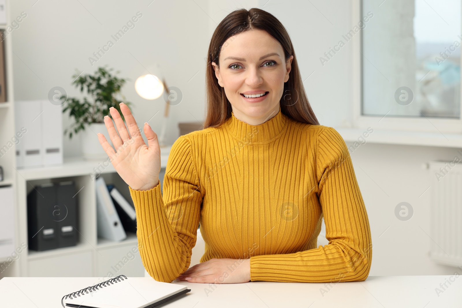 Photo of Woman waving hello during video chat at table in office, view from web camera