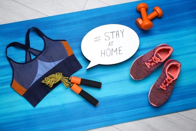 Photo of Sport equipment and speech bubble with hashtag STAY AT HOME on blue yoga mat, flat lay. Message to promote self-isolation during COVID‑19 pandemic