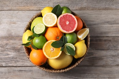 Photo of Different fresh citrus fruits and leaves in wicker basket on wooden table, top view