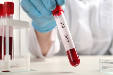 Photo of Scientist holding tube with blood sample and label STD Test at white table, closeup