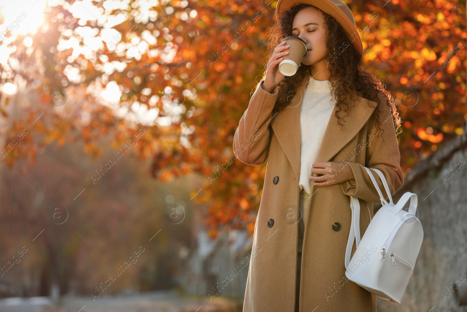 Photo of Beautiful African-American woman with stylish white backpack drinking coffee on city street. Space for text