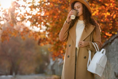 Beautiful African-American woman with stylish white backpack drinking coffee on city street. Space for text