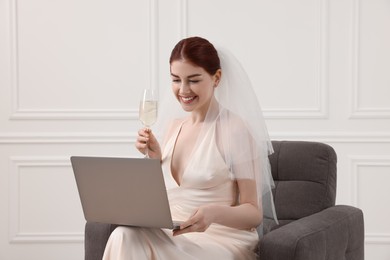 Photo of Happy bride with glass of sparkling wine and laptop indoors