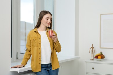 Beautiful young woman drinking delicious smoothie near window indoors. Space for text