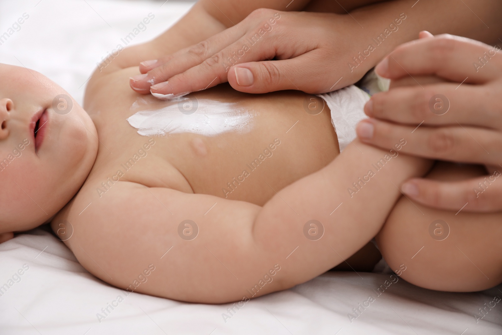 Photo of Mother applying moisturizing cream onto her little baby's skin on bed, closeup