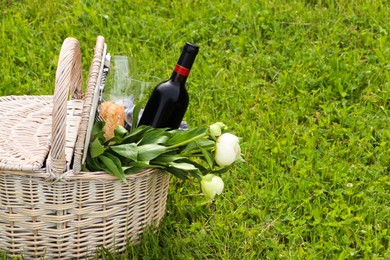 Picnic basket with wine, bread and flowers on green grass outdoors