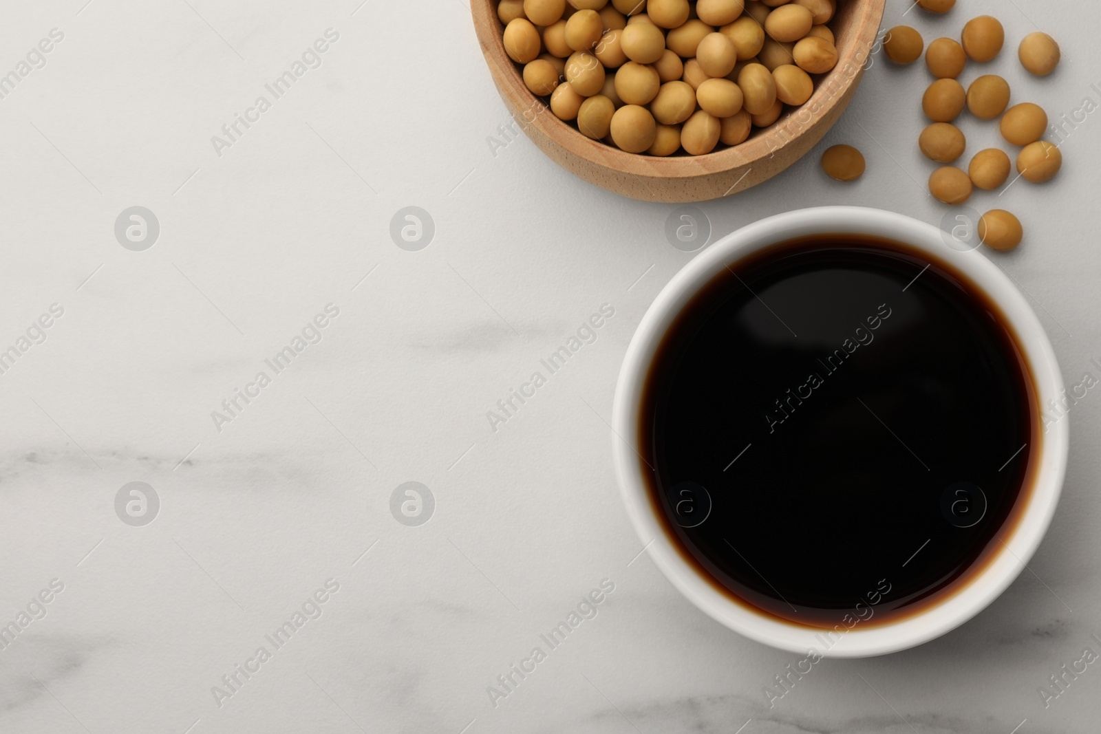 Photo of Soy sauce in bowl and soybeans on white marble table, flat lay. Space for text