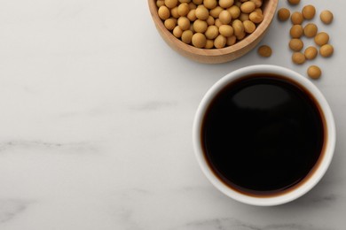 Soy sauce in bowl and soybeans on white marble table, flat lay. Space for text