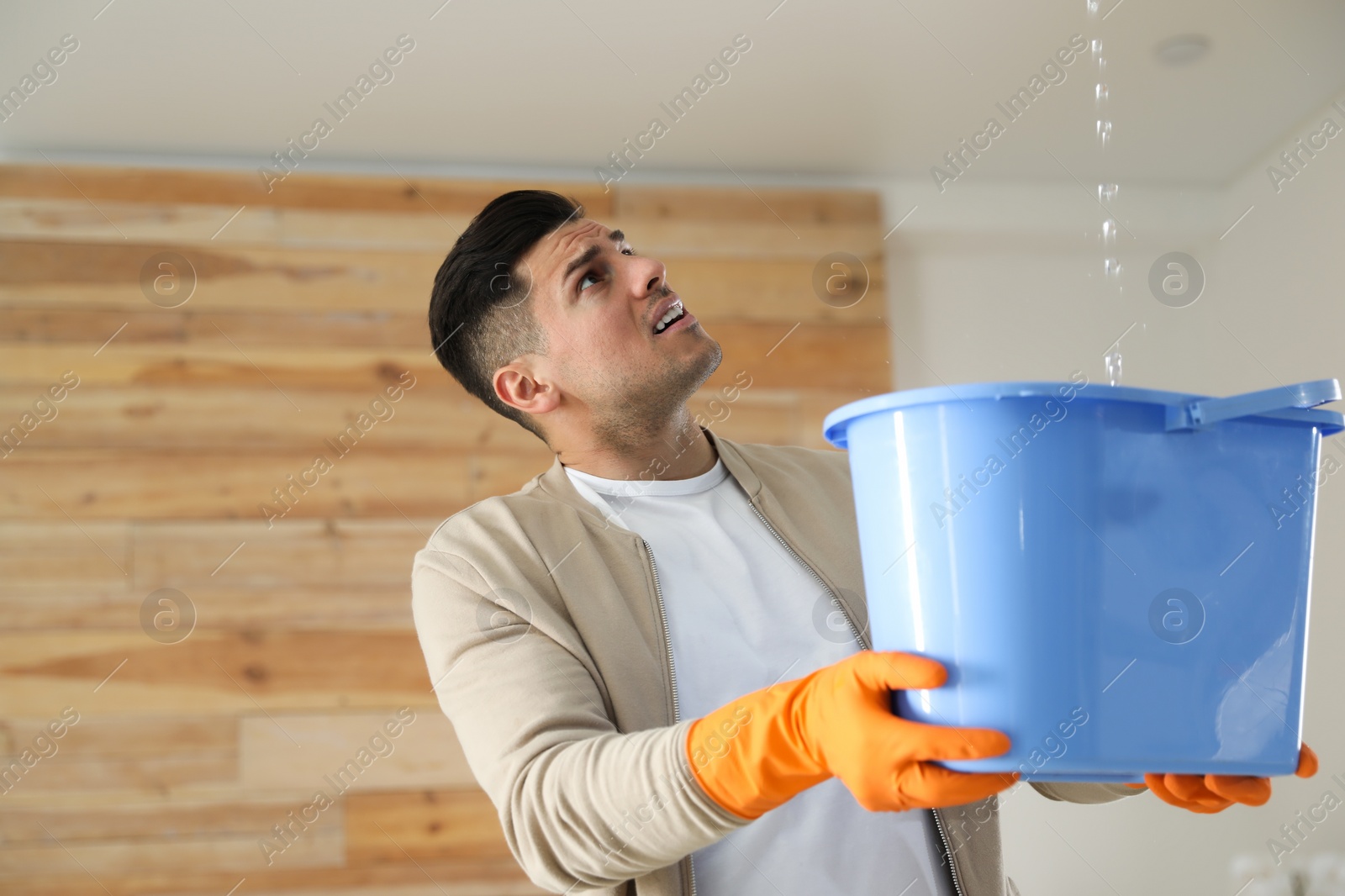 Photo of Emotional man collecting water leaking from ceiling indoors. Damaged roof