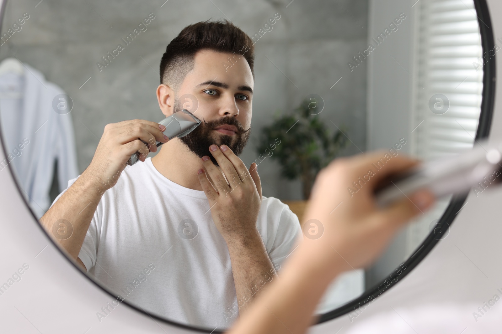 Photo of Handsome young man trimming beard near mirror in bathroom